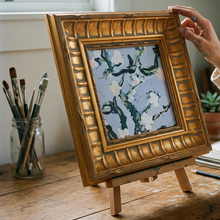 Load image into Gallery viewer, Artist holding Nineveh Floral Square wall art in an ornate gold frame on a studio desk easel with paintbrushes.